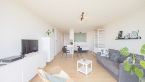 Un salon moderne avec un canapé gris, un fauteuil en osier, une petite table blanche et un téléviseur posé sur un meuble blanc.