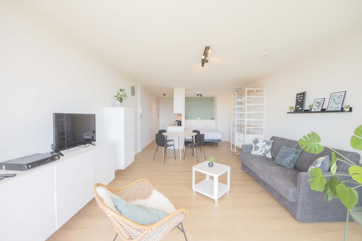 Un salon moderne avec un canapé gris, un fauteuil en osier, une petite table blanche et un téléviseur posé sur un meuble blanc.