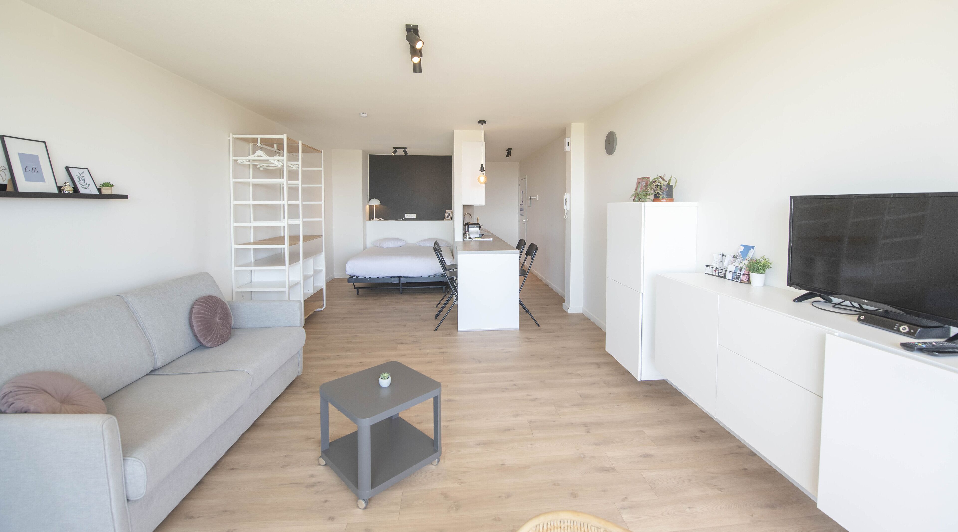 Een moderne, goed verlichte woonkamer met een grijze bank, een klein grijze tafel, een wit kastenmeubel, een televisie, en een keukenhoek met een koelkast en een magnetron.