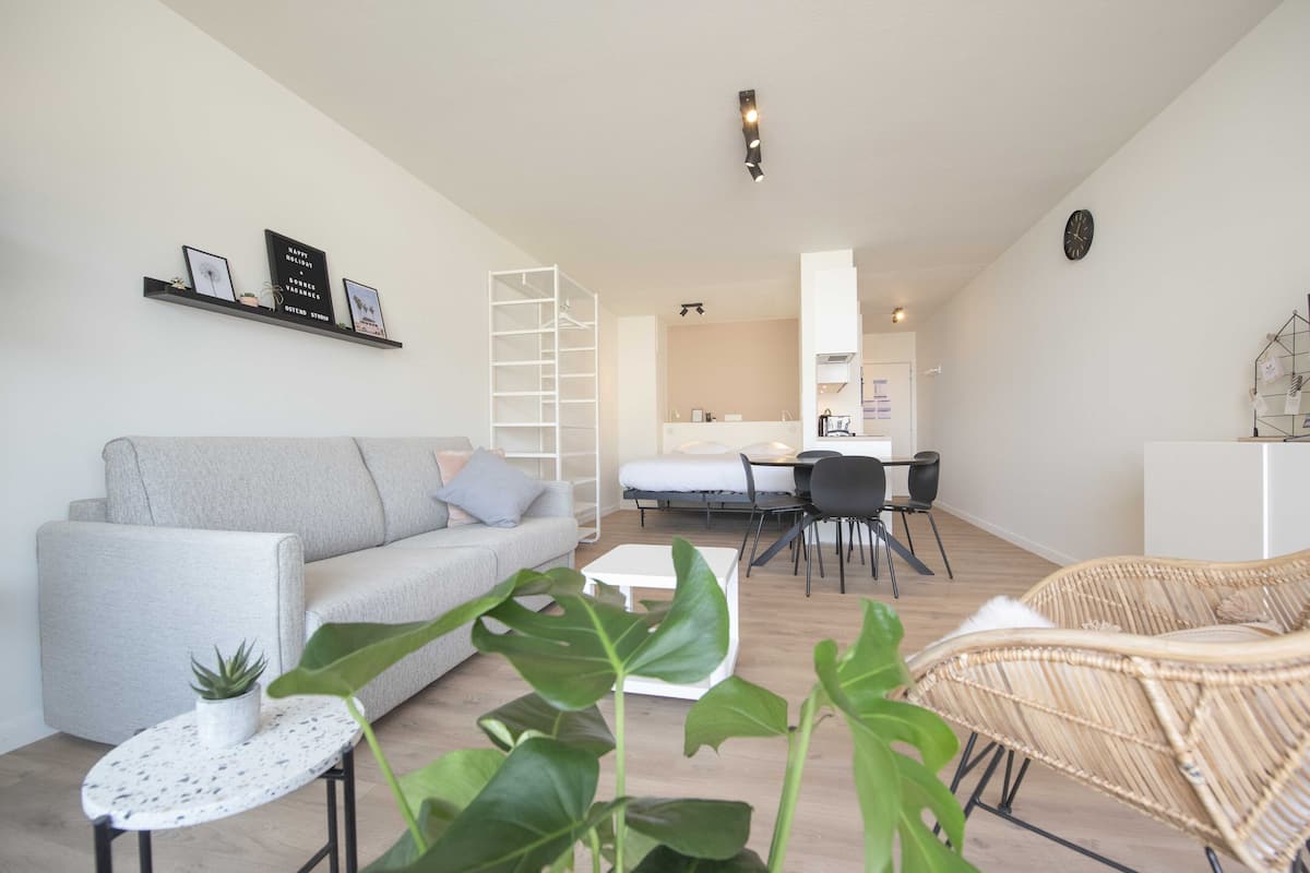 Un salon moderne avec un canapé gris, une table basse blanche et un fauteuil en osier. On y trouve également un coin repas avec une table et des chaises, et une cuisine à l’arrière-plan.