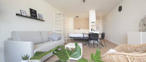 A modern living room with a grey sofa, a white coffee table, and a wicker chair. There is a dining area with a table and chairs, and a kitchen in the background.