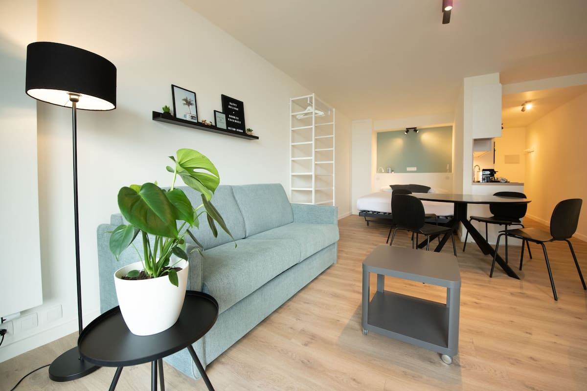 Un salon moderne avec un canapé bleu clair, un lampadaire noir, une plante en pot blanche, une table basse grise et un coin repas comprenant une table blanche et des chaises noires.