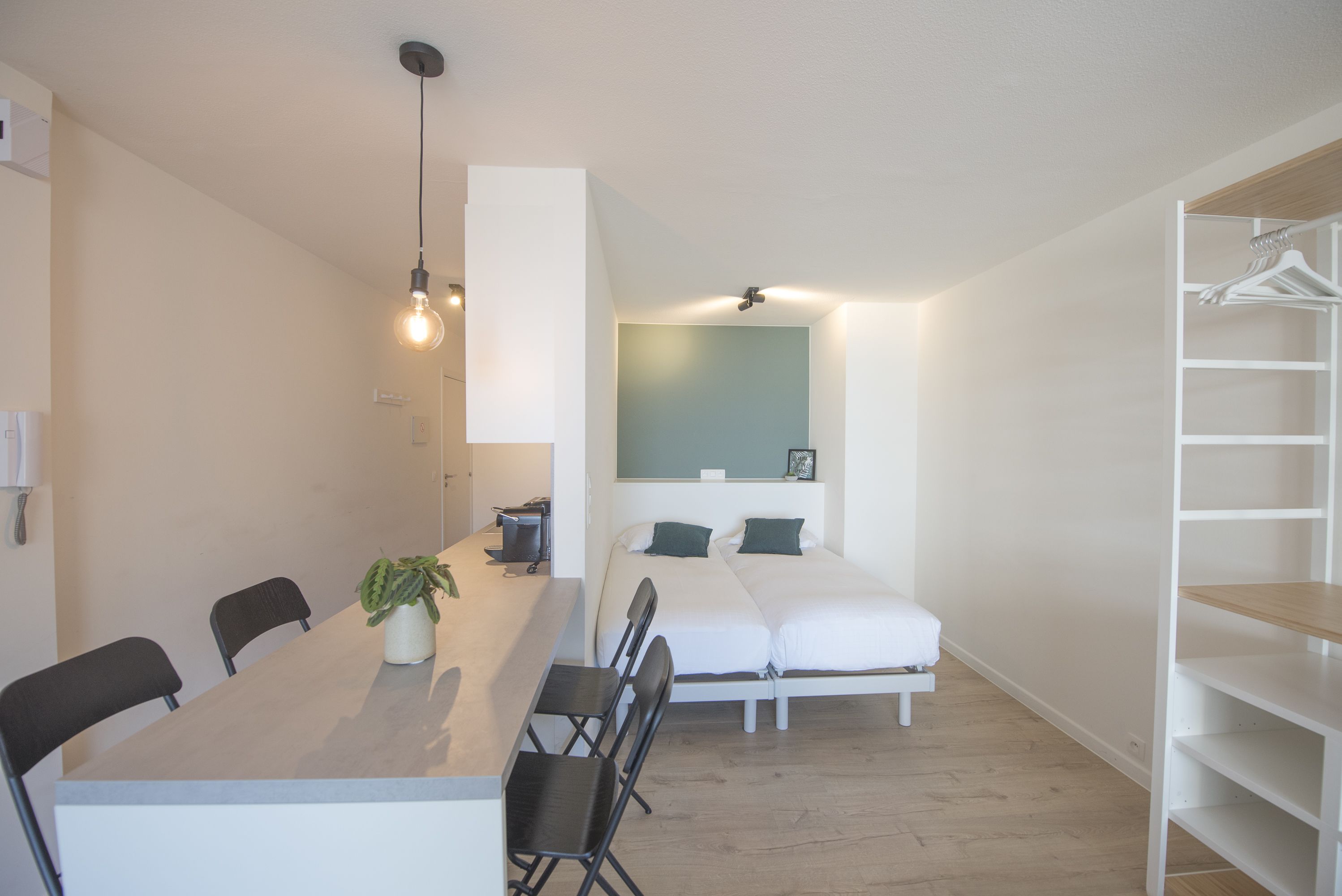 A compact room with a dining table, chairs, a small plant, a bed with two pillows, and a white wardrobe.