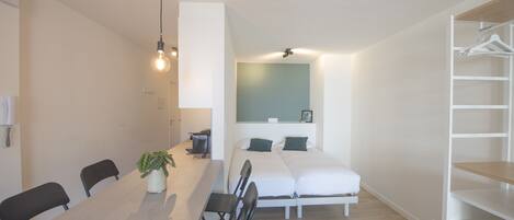 A compact room with a dining table, chairs, a small plant, a bed with two pillows, and a white wardrobe.