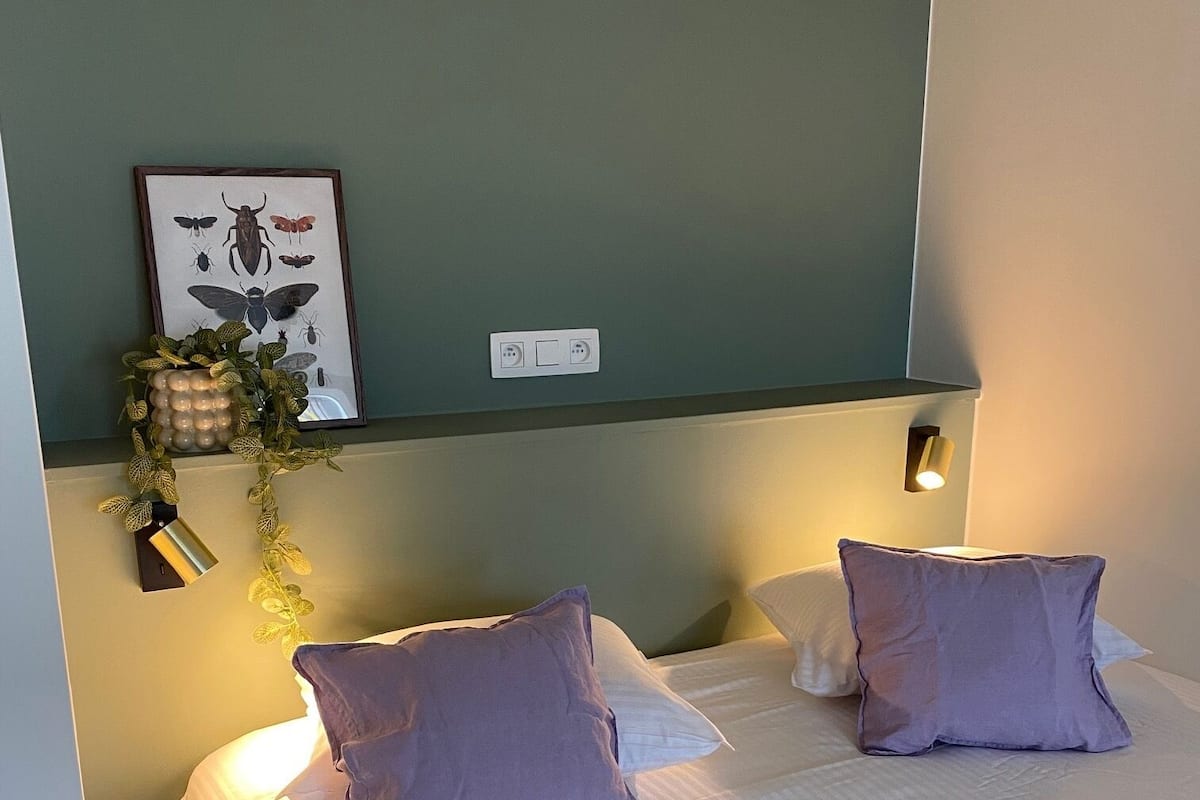 Un lit double avec du linge de lit blanc et des oreillers violets, un luminaire fixé au mur et une illustration d’insecte encadrée.