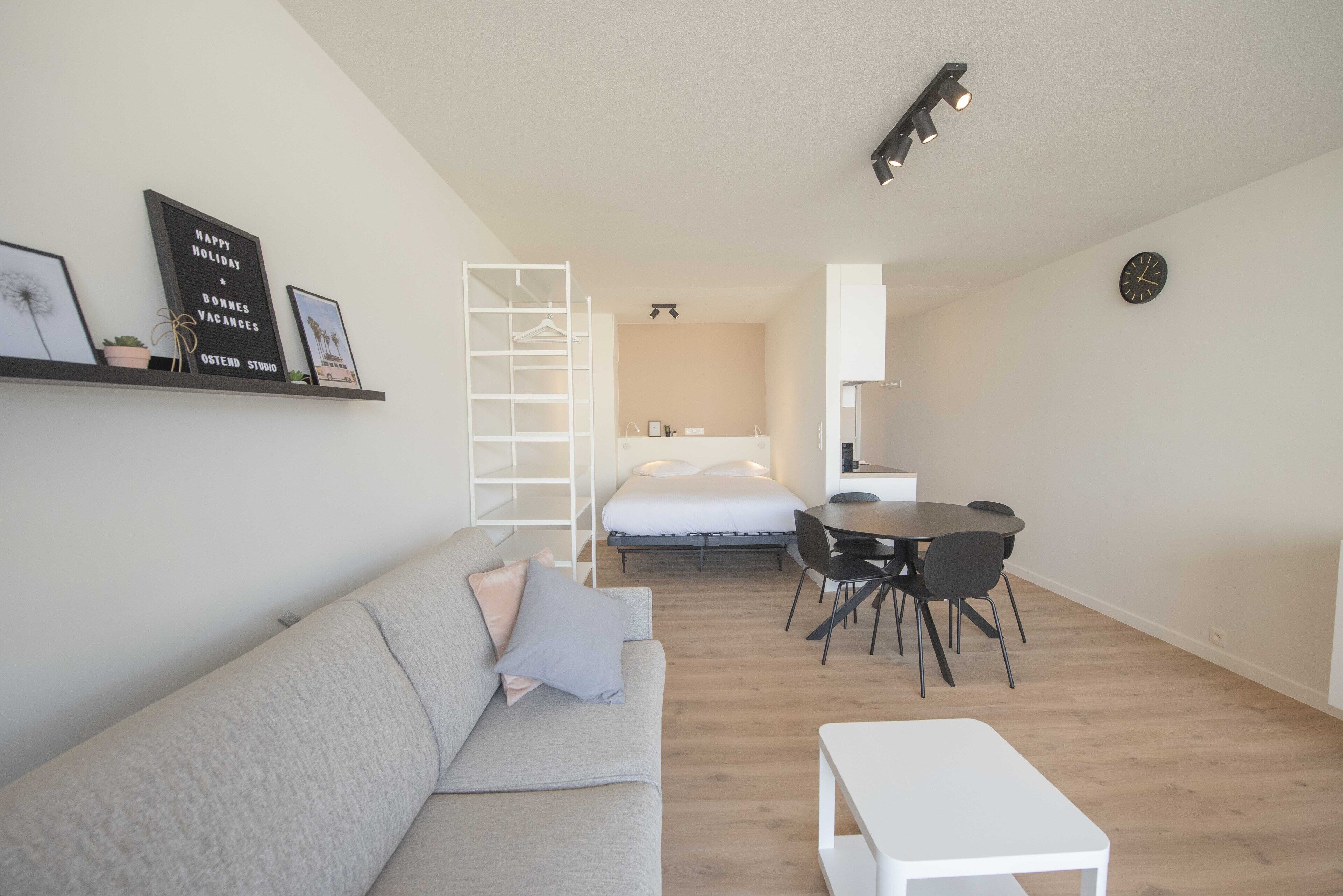 Ett modernt, väl upplyst rum med en grå soffa, ett litet vitt bord, en matplats med ett runt bord och stolar, en säng med en vit madrass och en vit garderob.