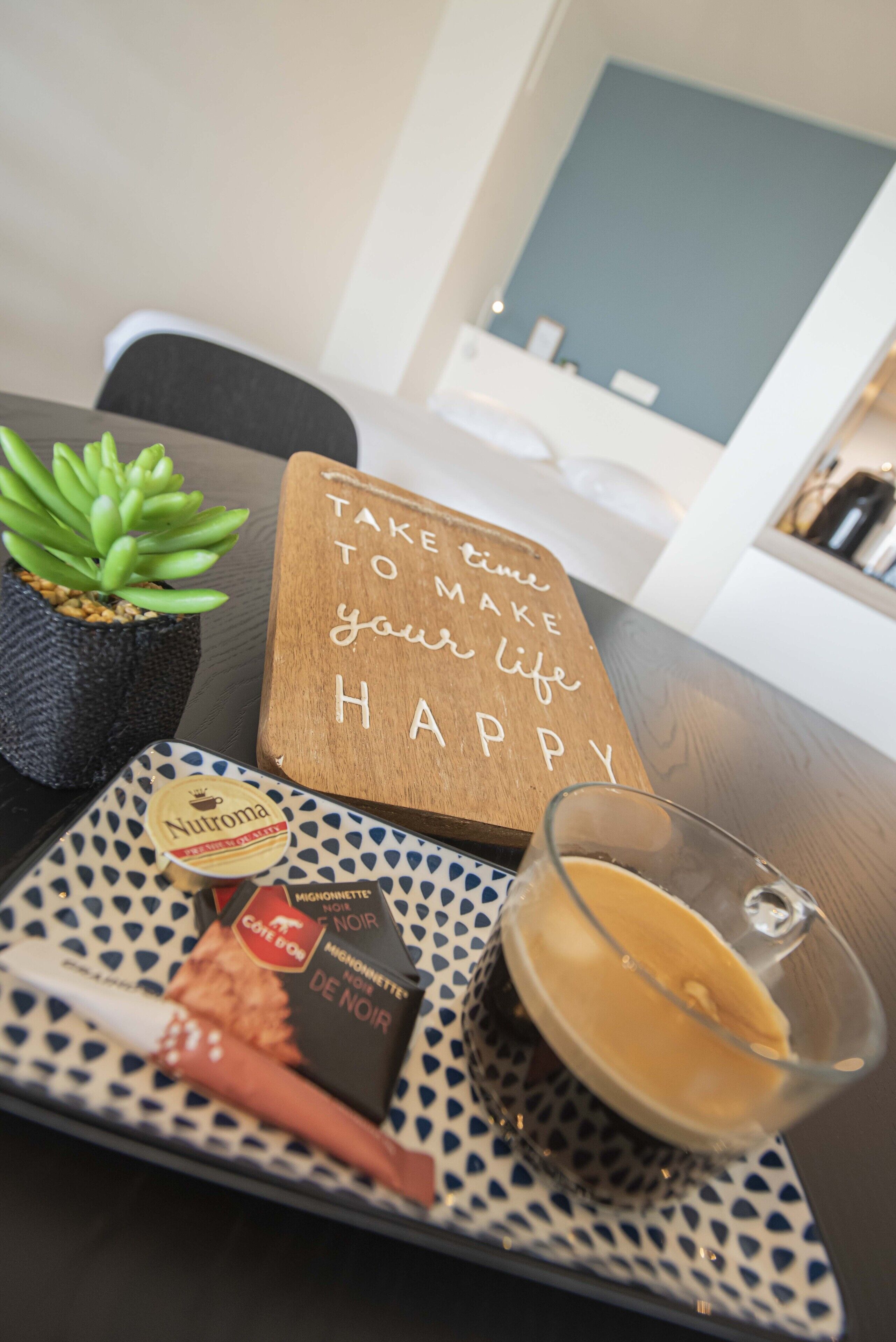 Et bord med et træskilt, en sukkulentplante, en bakke med en Nutroma chokoladebar, et glas kaffe og en flaske vin.