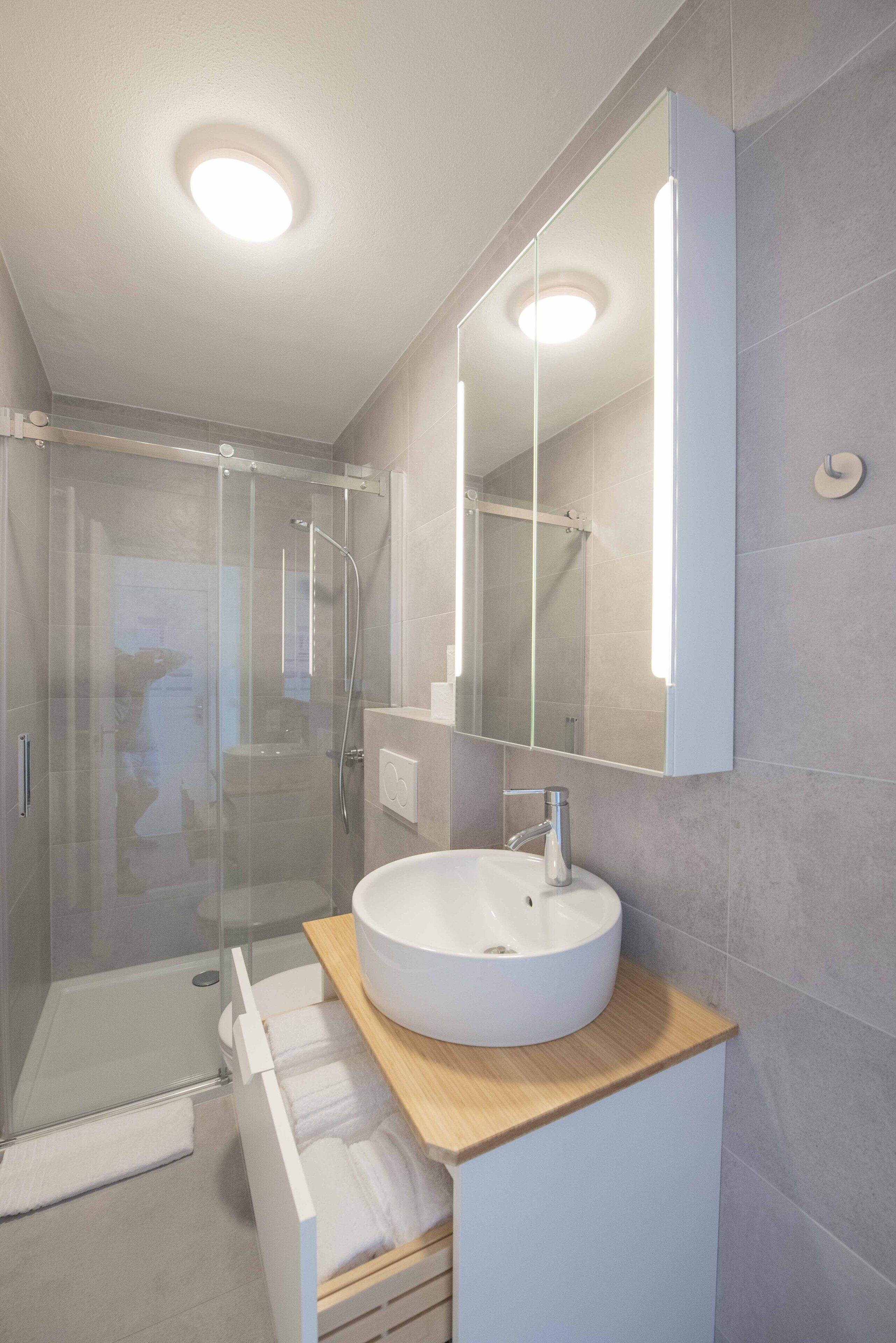 Een moderne badkamer met een glazen douchecabine, een wit wastafeltje en een grote spiegel.