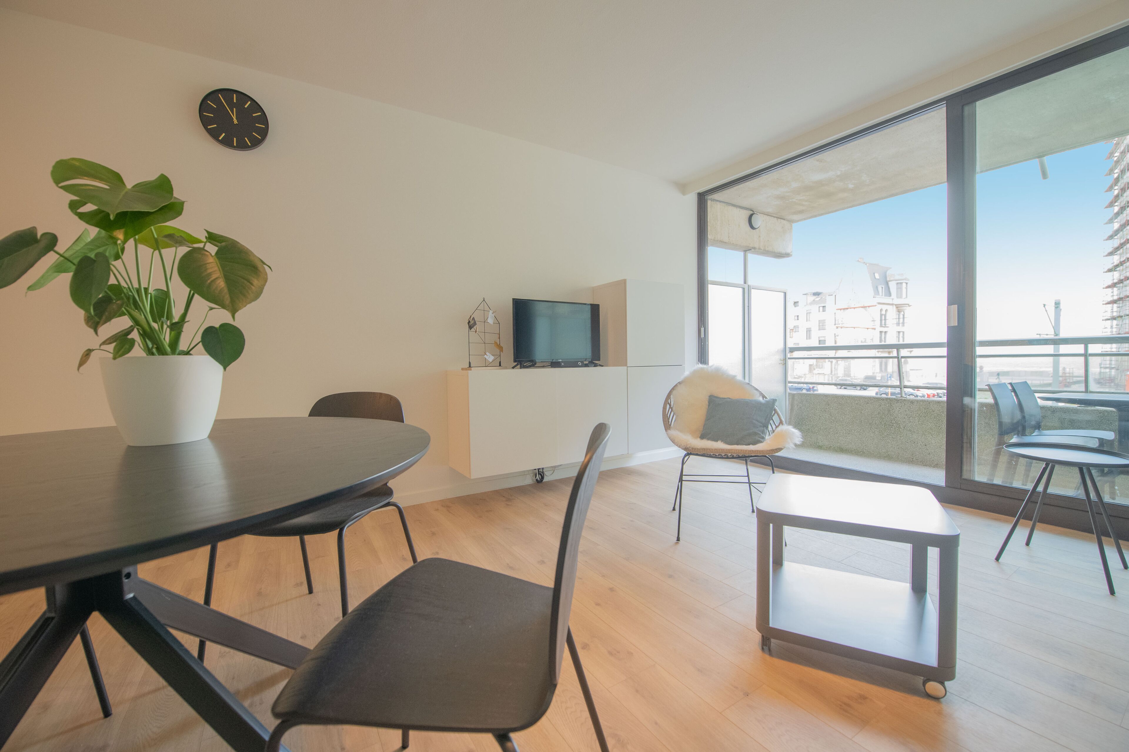 A modern living room with a dining table, chairs, a potted plant, a TV, and a large window offering a view of buildings.