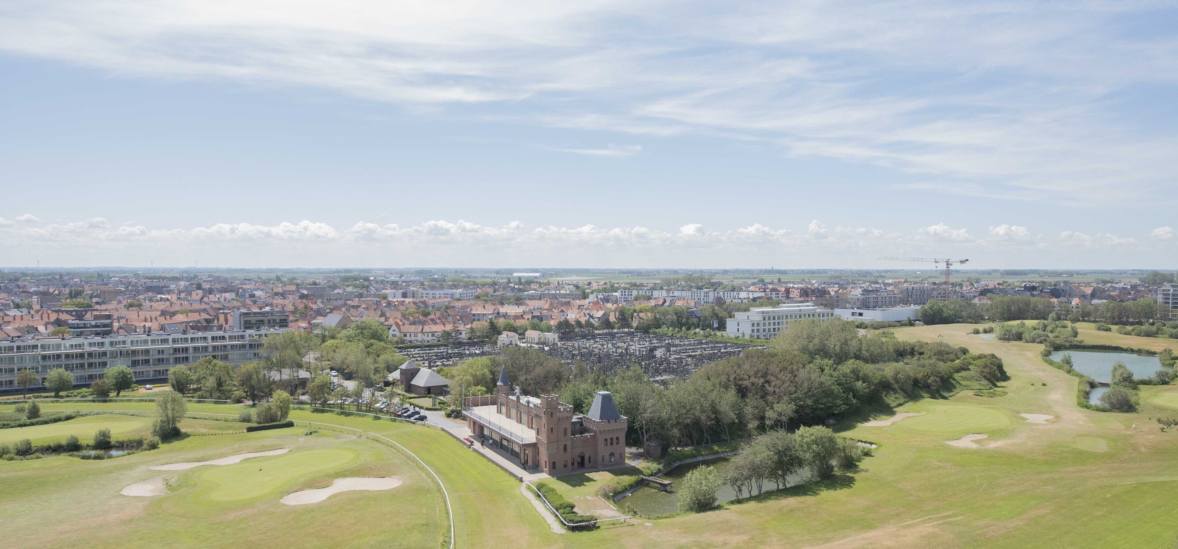 Vista aerea di un paesaggio urbano con un campo da golf, un edificio simile a un castello e un grande edificio moderno.