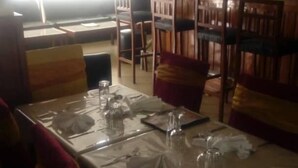 Ein Essbereich mit einem für zwei Personen gedecktem Tisch, eine Bar mit einer Person hinter dem Tresen und ein Fenster, das natürliches Licht hereinlässt.