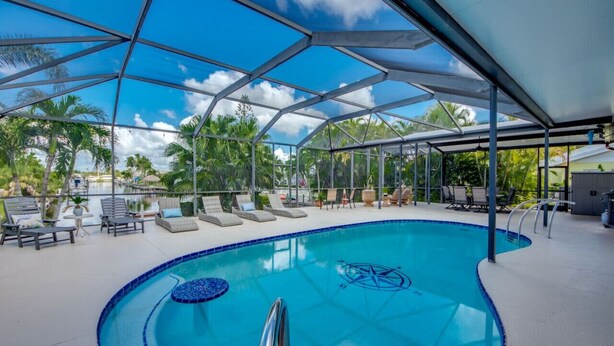 A pool area with lounge chairs, a covered seating area, and a clear view of palm trees. - Coastal Heaven: Huge Lanai-Heated Pool-Canal w Private Dock-River Views (Cape Coral)