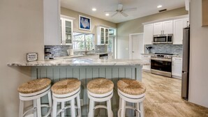 A kitchen with white cabinets, a marble countertop, a stainless steel refrigerator, and a ceiling fan. - Coastal Heaven: Huge Lanai-Heated Pool-Canal w Private Dock-River Views (Cape Coral)