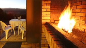 A patio with a table and chairs, a brick fireplace with a fire, and a view of distant hills.