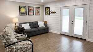 A living room with a grey sofa, patterned armchairs, a ceiling fan, and framed posters on the wall. - The Cottage on College - Pets & Parking (Brandon)