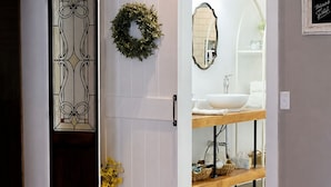 A room with a sliding door leading to a bathroom. The bathroom has a white sink, a mirror, and a wooden bench. There is a wreath on the wall and a vase with flowers. - The Cottage on College - Pets & Parking (Brandon)