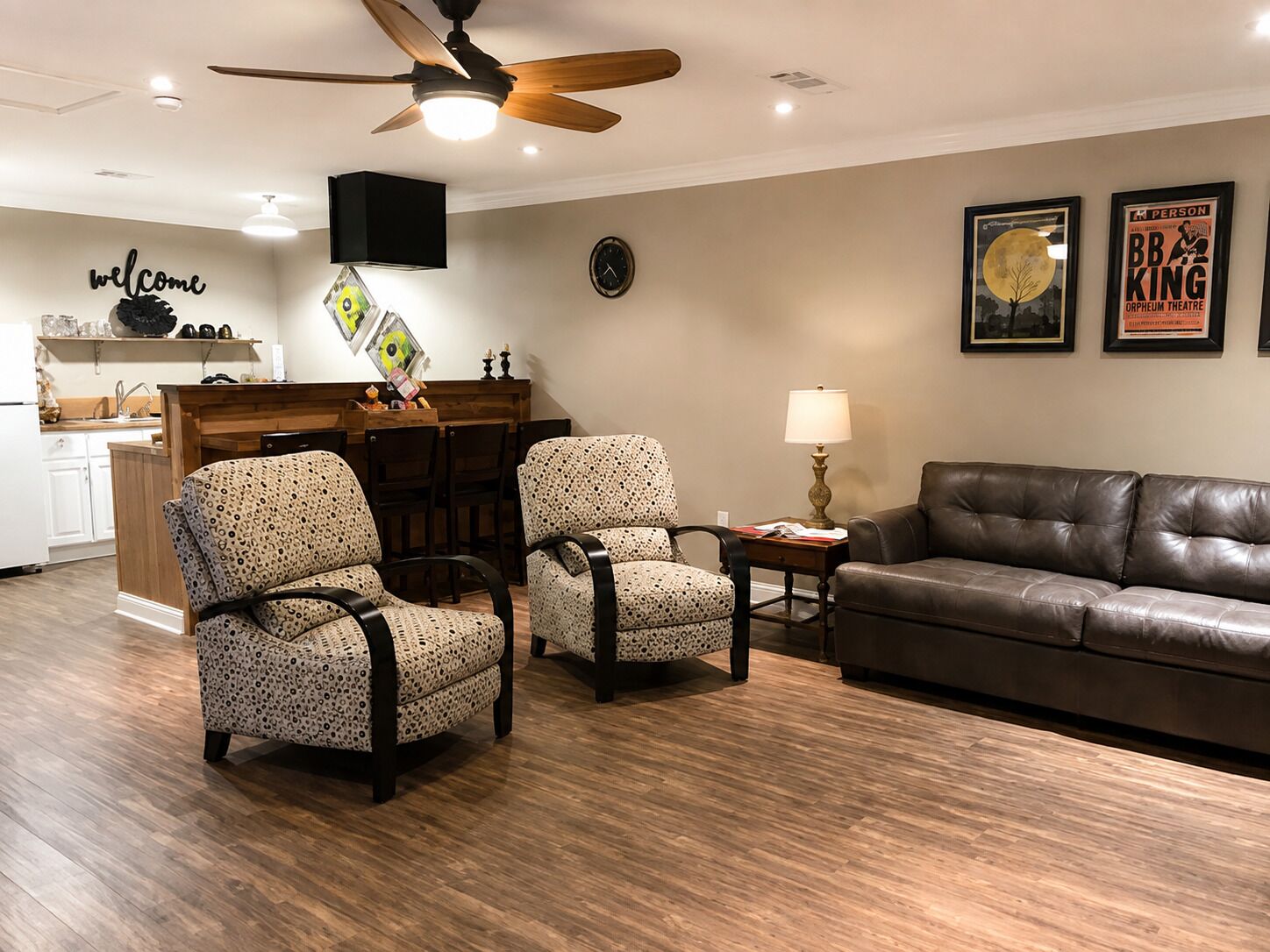 A living room with a brown leather sofa, two patterned armchairs, a wooden coffee table, a ceiling fan, a wall clock, and framed posters.