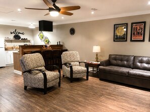 A living room with a brown leather sofa, two patterned armchairs, a wooden coffee table, a ceiling fan, a wall clock, and framed posters. - The Cottage on College - Pets & Parking (Brandon)