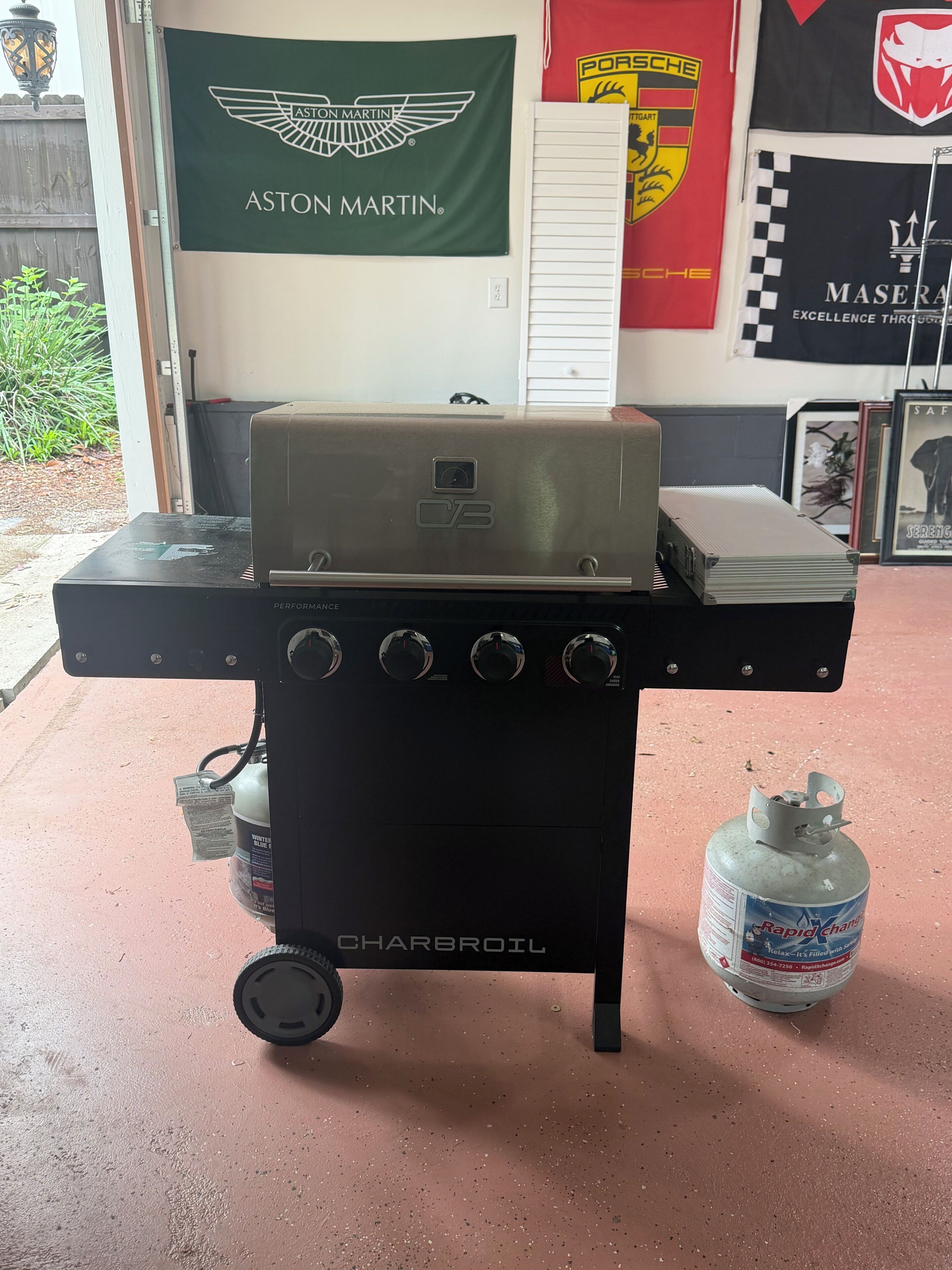 A Charbroil grill with a gas tank and propane cylinder, set on a concrete floor with a garage door and banners in the background.