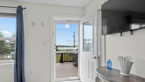 A room with a wooden floor, a white door leading to a balcony, a flat-screen TV mounted on the wall, and a small table with a blue bottle and a white vase. - Beautiful View of Rustico Bay (North Rustico)