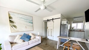 A modern kitchen with a dining area, a white sofa with patterned cushions, a ceiling fan, and a beach-themed painting. - SALT by the Sea - Unit 2 (Port Macquarie)