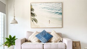 A living room with a beige sofa, a large beach-themed painting, a hanging light fixture, and a potted plant. - SALT by the Sea - Unit 2 (Port Macquarie)