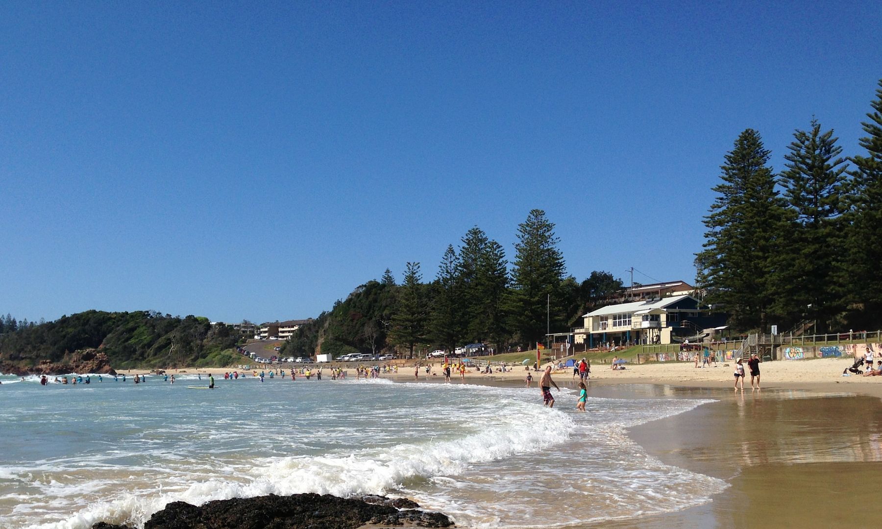A beach with clear skies, waves, and people. There are trees and buildings in the background.