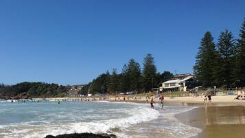 A beach with clear skies, waves, and people. There are trees and buildings in the background.