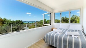 Een slaapkamer met een groot raam, een bed met gestreept beddengoed en uitzicht op groen en een verafgelegen landschap.
