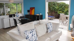 Een moderne woonkamer met een witte leren bank, een bureau met een computer, en uitzicht op een tuin door grote ramen.