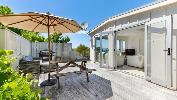 Een houten terras met een picknicktafel en parasol, een barbecue en een glazen ruimte met een televisie.