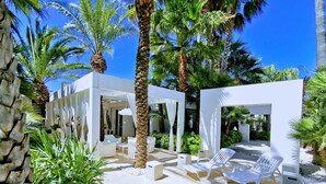 Un'area lounge esterna moderna con mobili bianchi, palme e un cielo azzurro intenso.