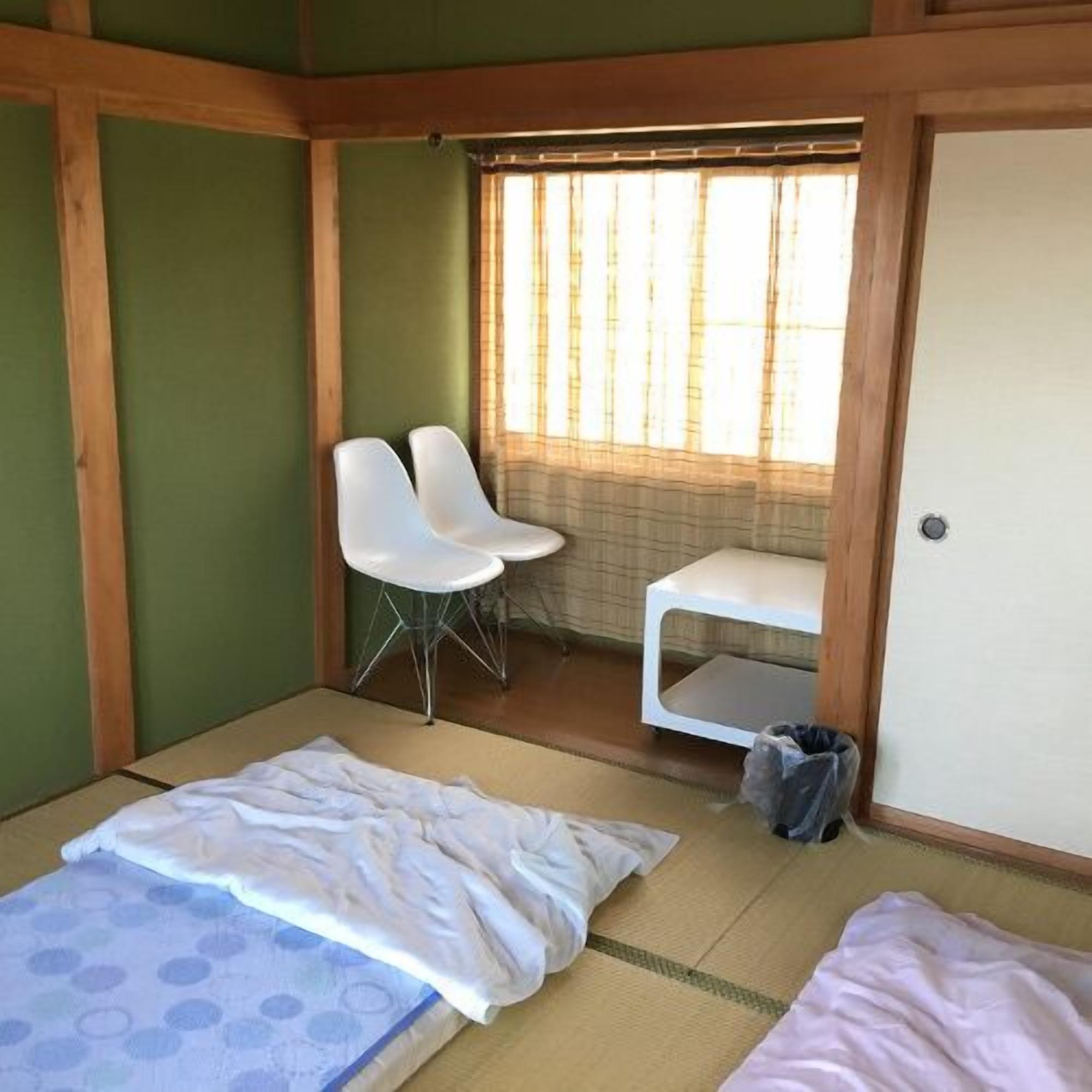 Una habitació amb sòl de tatami, un llit amb col·cha de disseny blau i blanc, dues cadires blanques, una taula petita blanca i una finestra amb persianes verticals.