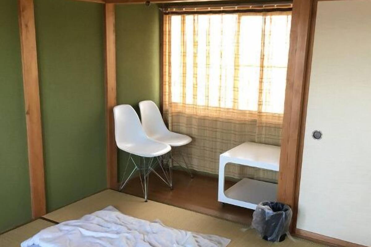 A room with tatami flooring, a bed with a blue and white patterned bedspread, two white chairs, a small white table, and a window with vertical blinds.