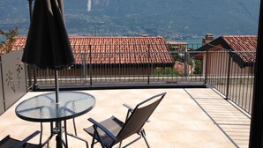 A balcony with a glass table and chairs, a black umbrella, and a view of mountains and a lake.