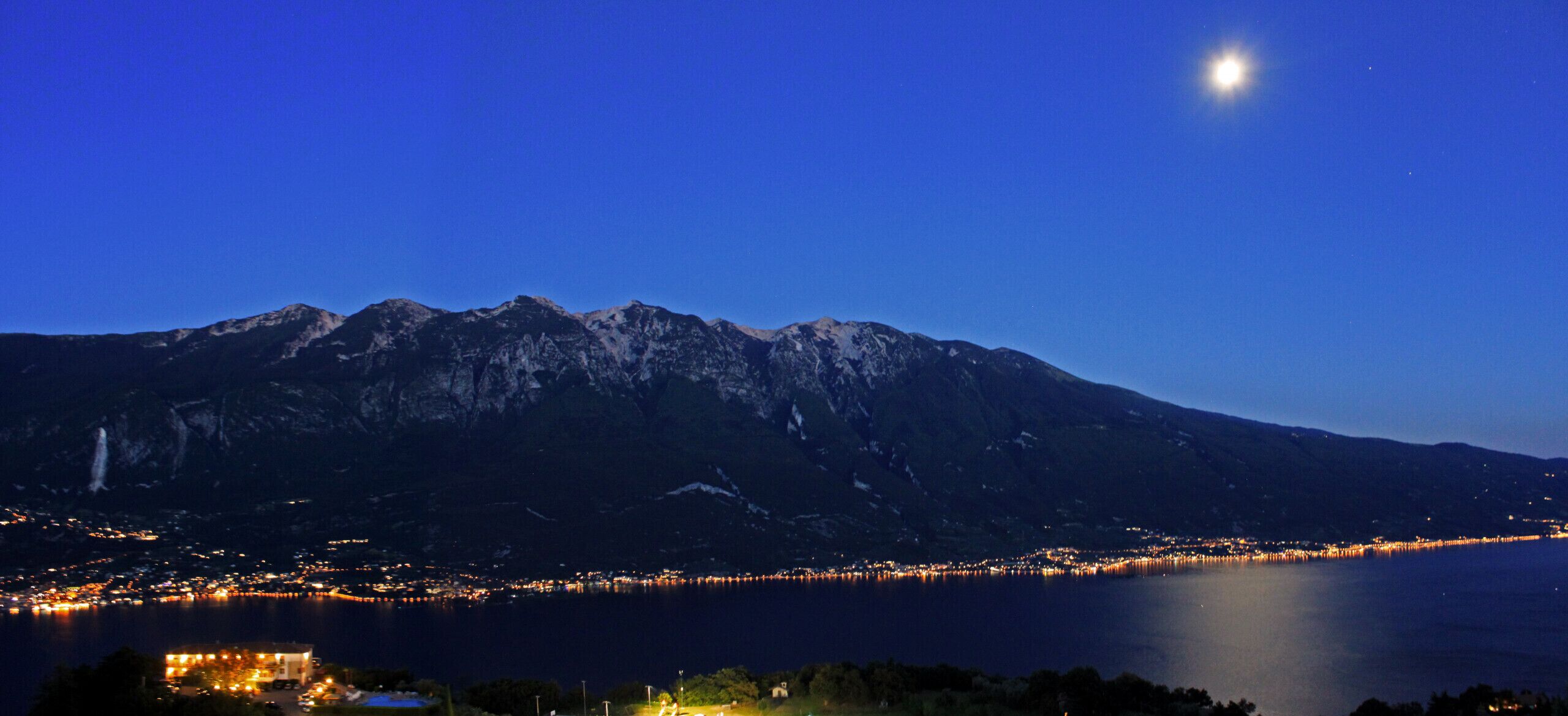 A mountain range at dusk with a lake in the foreground and a town along the shoreline.