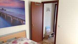A bedroom with a bed, a painting of a pier, a wooden door, and a small shelf.