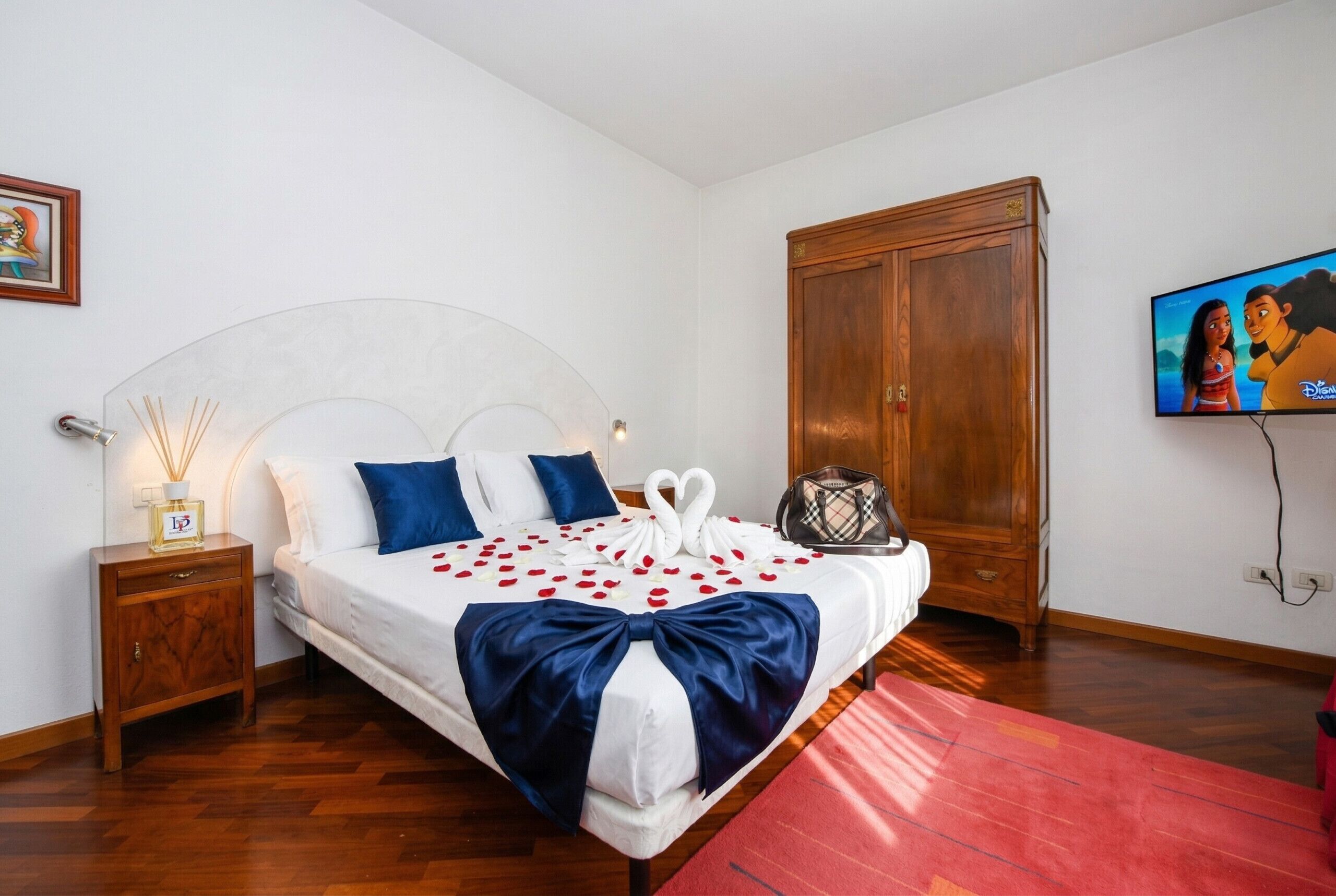 A neatly made bed with a bow, a wooden wardrobe, a television, and a small table with a bottle and candle.