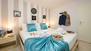 Un lit bien fait, recouvert d’une couverture bleue, une table de chevet avec une lampe et une horloge murale.