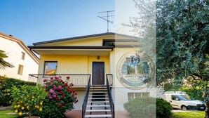 Un bâtiment jaune avec un escalier menant à l’entrée, entouré de verdure et avec une voiture garée à proximité.