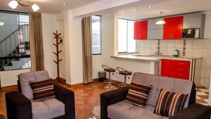 A modern living room with two sofas and a coffee table, a kitchen with red cabinets and a microwave, and a staircase leading to an upper floor. - Mayte Apartment (Cusco)