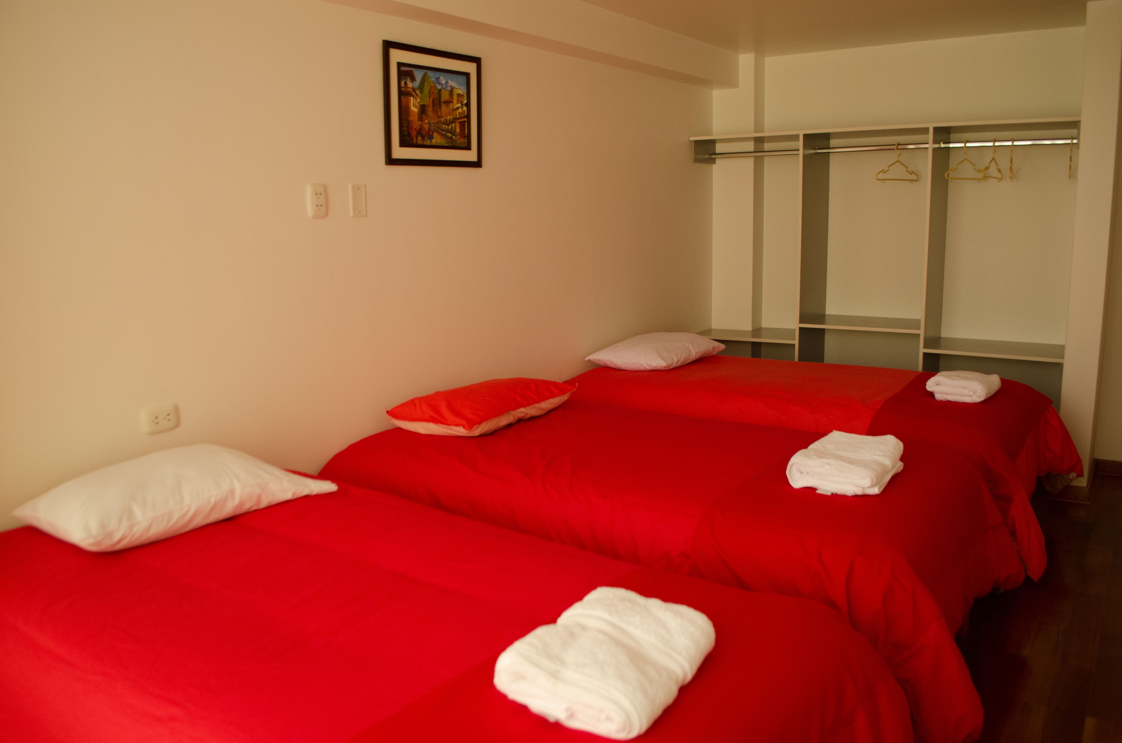 A hotel room with two beds, red bedding, white pillows, a painting on the wall, and a wardrobe with hangers.