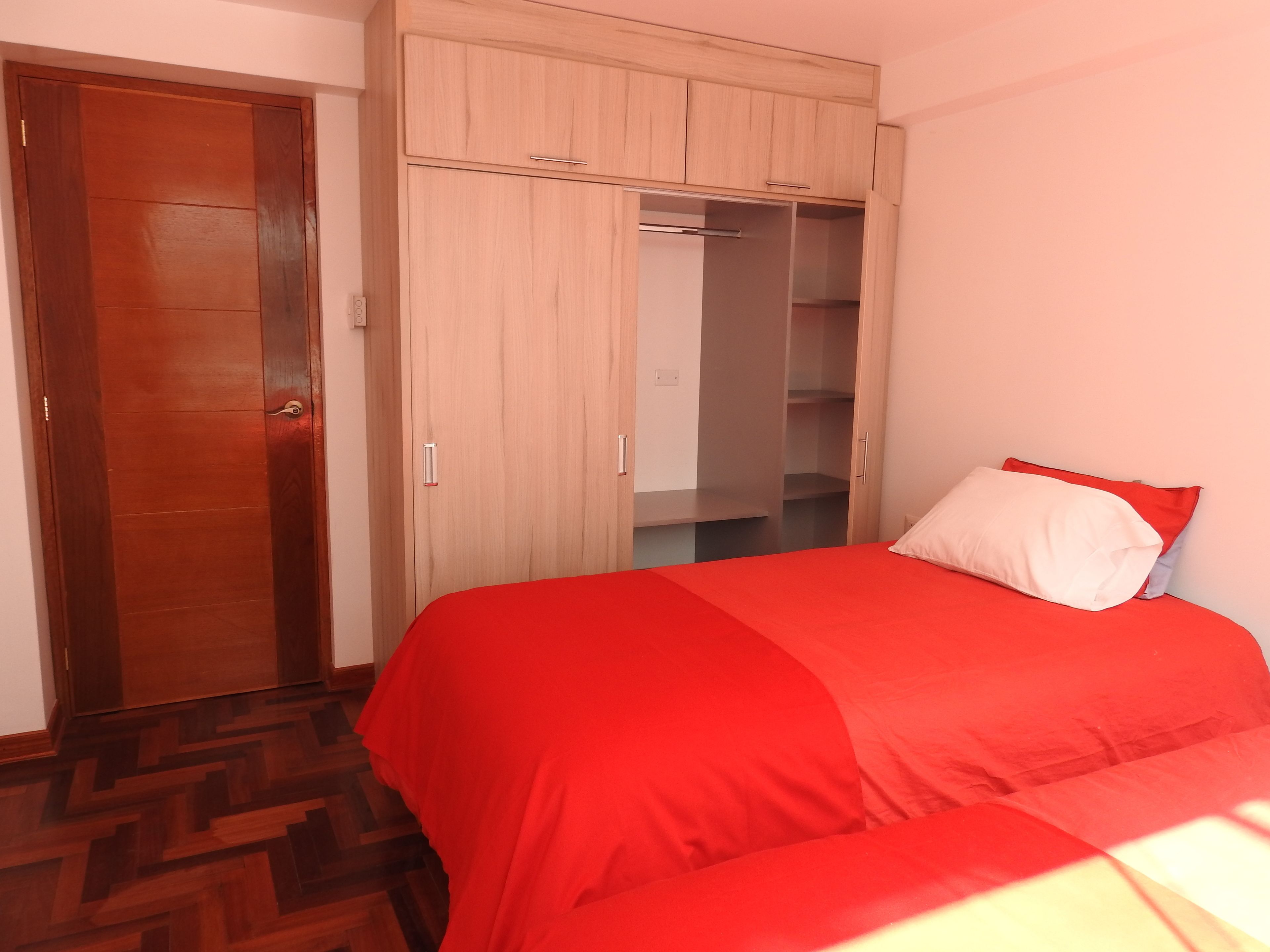 A bedroom with a wooden wardrobe, a bed with red bedding, and a herringbone-patterned floor.