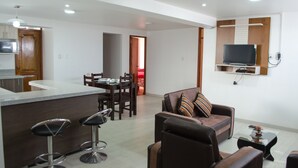 A modern living room with a sofa, a coffee table, and a TV mounted on the wall. There is a dining area with a table and chairs, and a kitchen area with a counter and stools. - Mayte Apartment (Cusco)