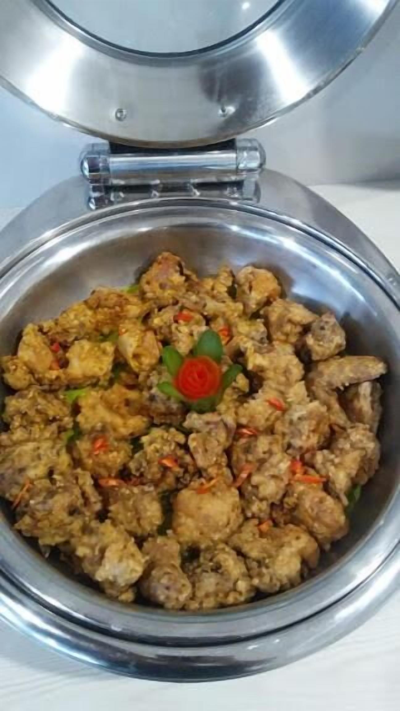 A stainless steel bowl with a lid, containing a dish with chunks of meat and vegetables, garnished with a red chili and green leaves.