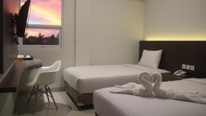 Ein Hotelzimmer mit zwei Betten, einem Schreibtisch mit Stuhl, einem an der Wand montierten Fernseher und Blick auf den Sonnenuntergang durch das Fenster.