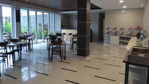 Eine moderne Hotelhalle mit sauber gefliestem Boden und einer zentralen Säule. Esstische und Stühle sind für Speisen arrangiert, und eine Küchenzeile mit Geräten und Geschirr ist sichtbar.