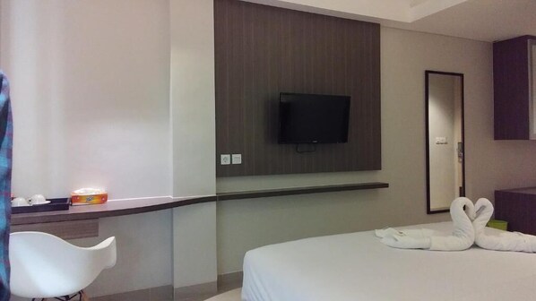 A hotel room with a bed, a TV mounted on the wall, a mirror, and a small desk with a chair. - Emerald Hotel (Timika)