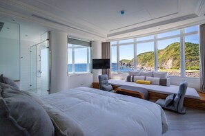 Una habitación de hotel moderna con una cama grande, una zona de estar con vistas al mar y un box de ducha de vidrio.
