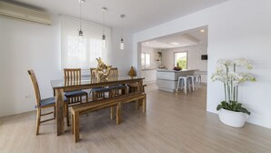 Een ruime woonkamer met een houten eettafel, stoelen en een bank. Achtergrond is een keuken met witte kasten en een bar.